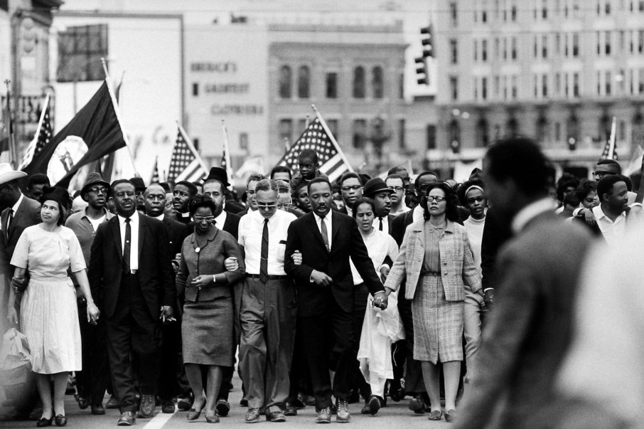 Martin Luther King leads a large crowd of African American men, women and children filling the street as they march towards the camera. They are dressed in formal 60s clothing, singing or chanting, with arms linked or hodling hands. A few people further back in the crowd are carrying American flags.