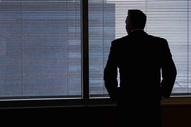 A man in a suit looks out of an office building window through venetian blinds at another office building