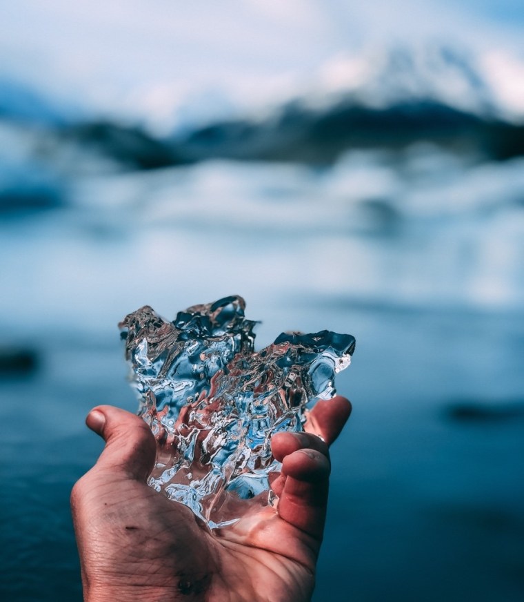 A hand holds a rough, asymmetrical piece of ice which reflects the colors of a blurred snowy mountain landscape in the background.
