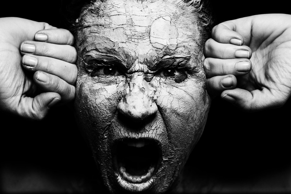A person is screaming. The face is covered with cracking mud, and the person's fists are pressing in one each side of their head