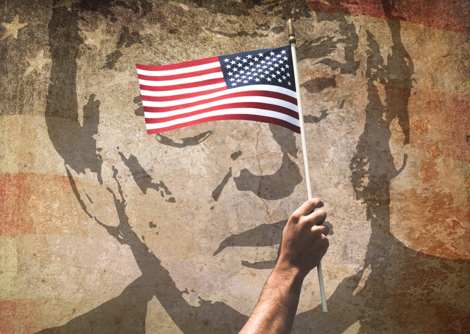In front of a faded wall mural of Donald Trump with an American flag background, a real hand holds up a real flag