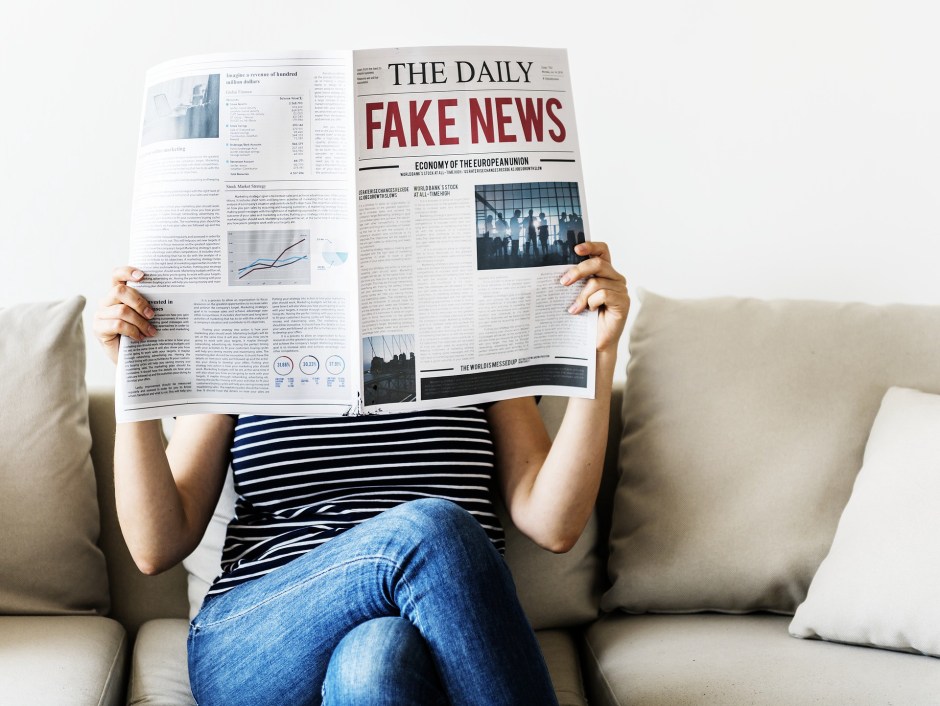 A woman sits on a couch reading a newspaper with the huge red headline 'FAKE NEWS'