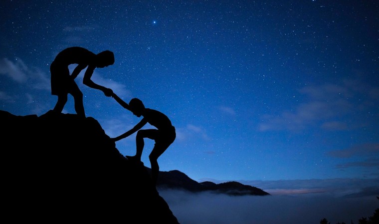 On a rocky mountain top at dusk or dawn, one boy helps another climb the last steps to the top