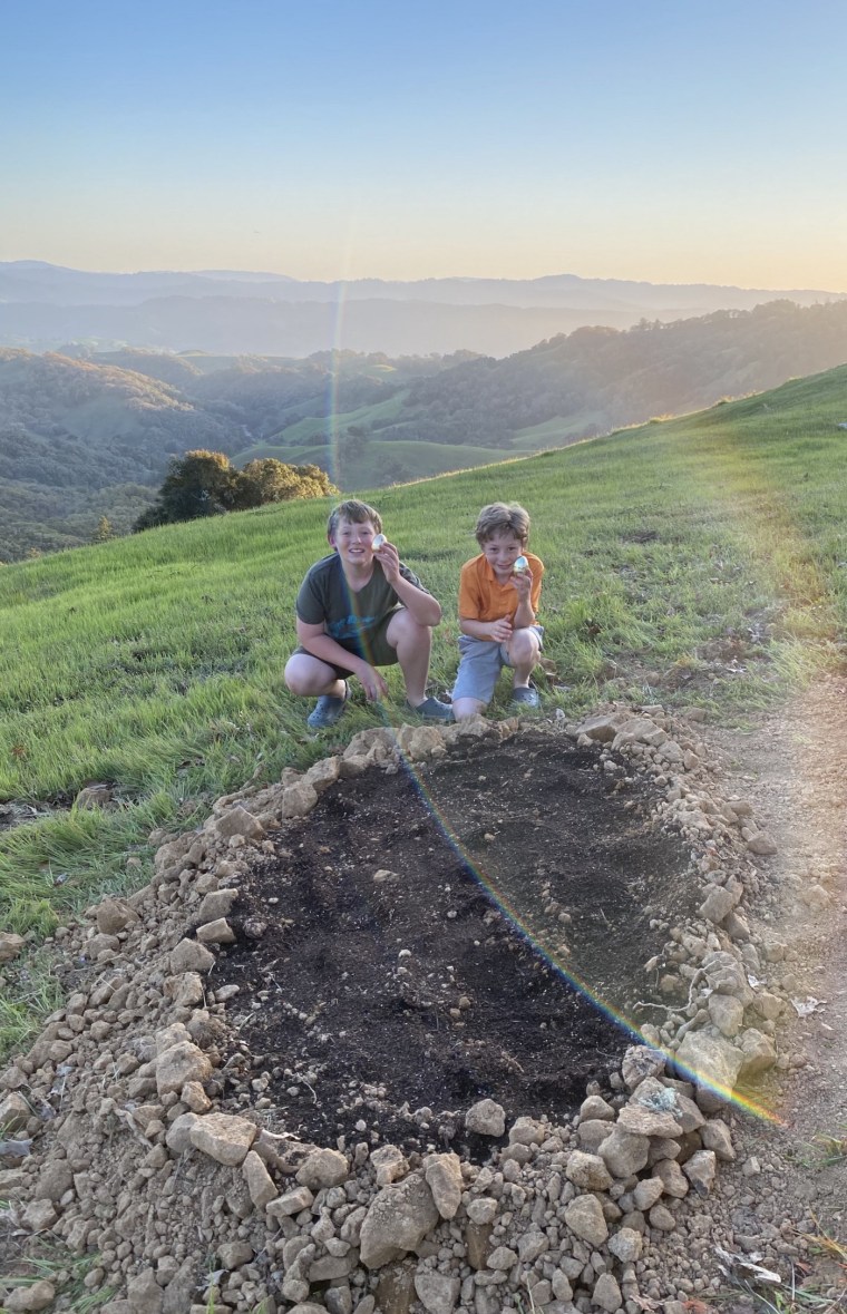 Two grinning boys wearing t-shirts and shorts, squatting at the edge of an oval patch of freshly turned earth surrounded by a border of stones and gravel, display their Easter eggs to the camera.