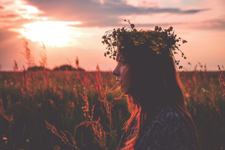 A woman stands in a field with the early evening sun on her face, wearing a wreath of grasses and small wildflowers