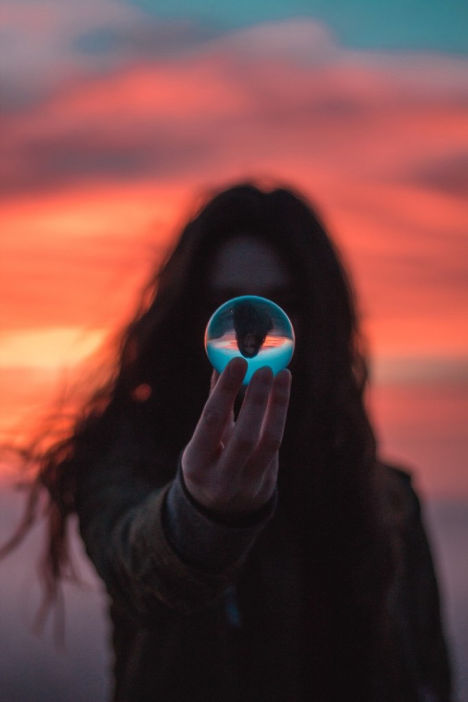 A woman silhouetted against a pink streaked sky stares into a small crystal ball that she extends out in front of herself with one hand. She is reflected upside down within the ball.